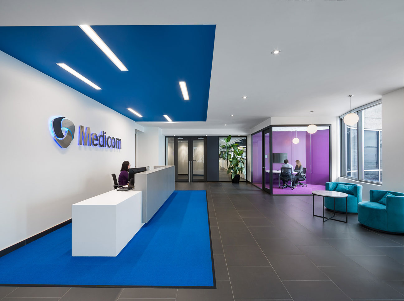 White reception desk on blue carpet with blue recessed ceiling, Medicom logo on wall and glass cube meeting area  finished in purple.