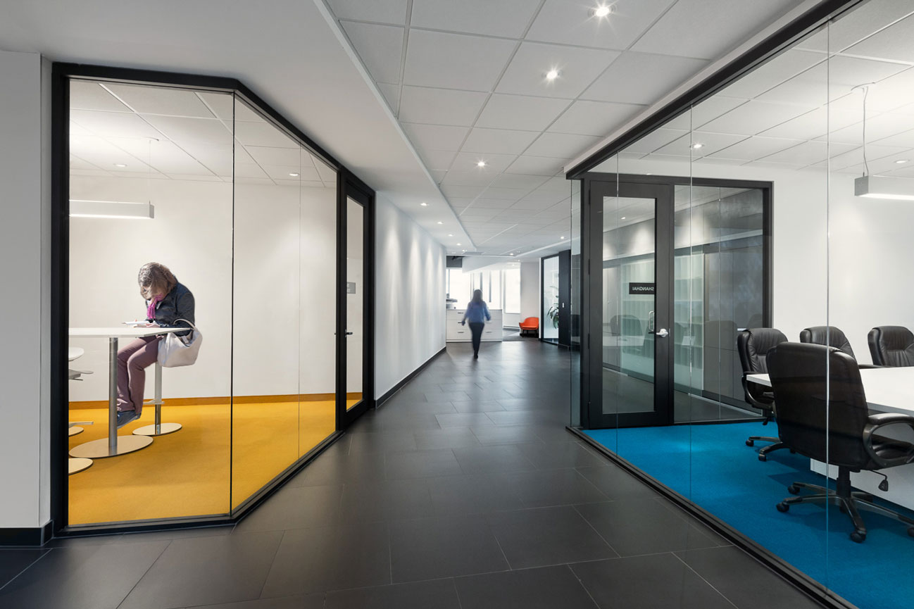 Corridor with glass cube meeting rooms on left and right, left with turquoise carpet, right with yellow wall and carpet.