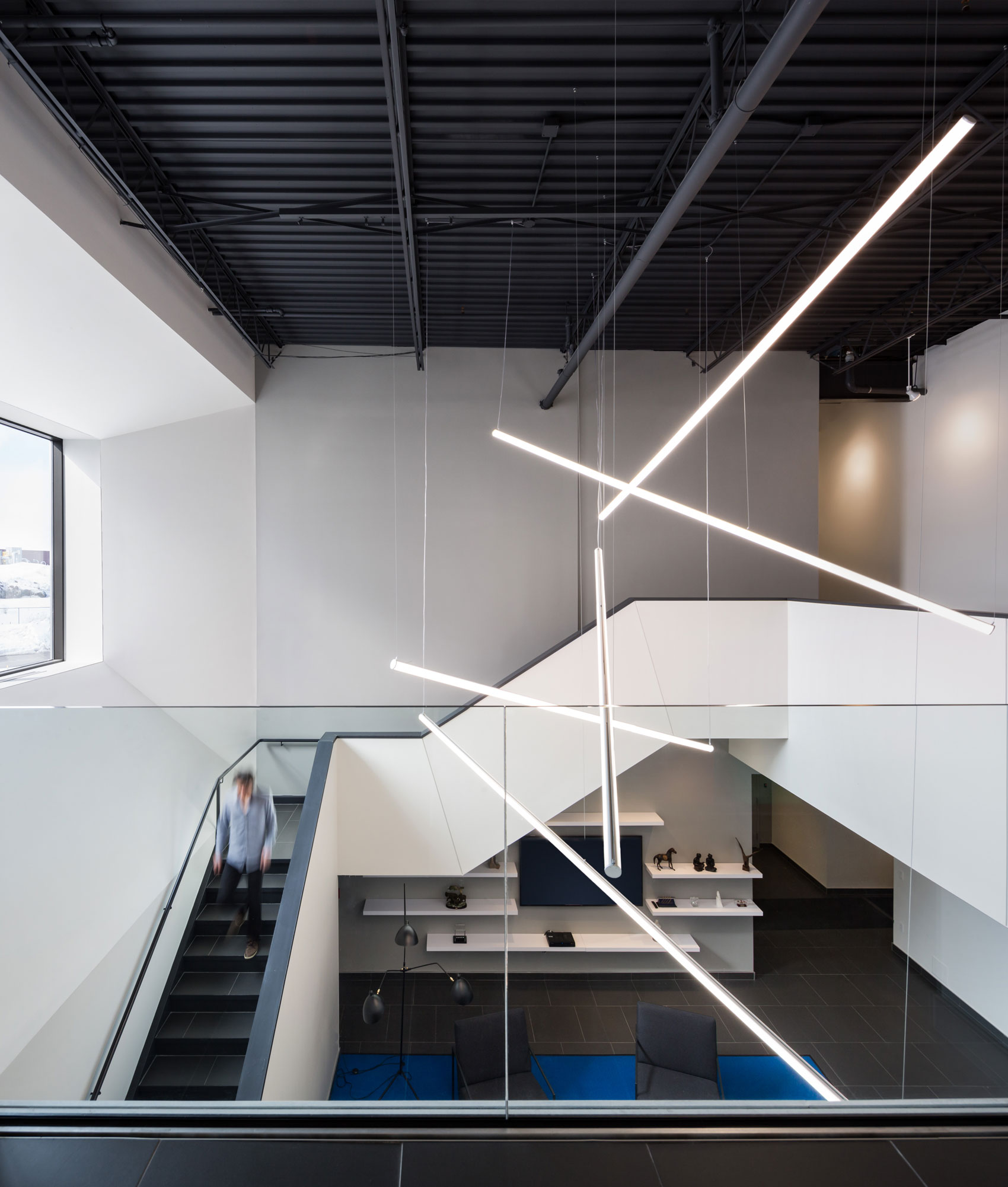 White mezzanine and stair with low level partition guard rail, angled suspended strip lights and blue carpeted lounge area below.