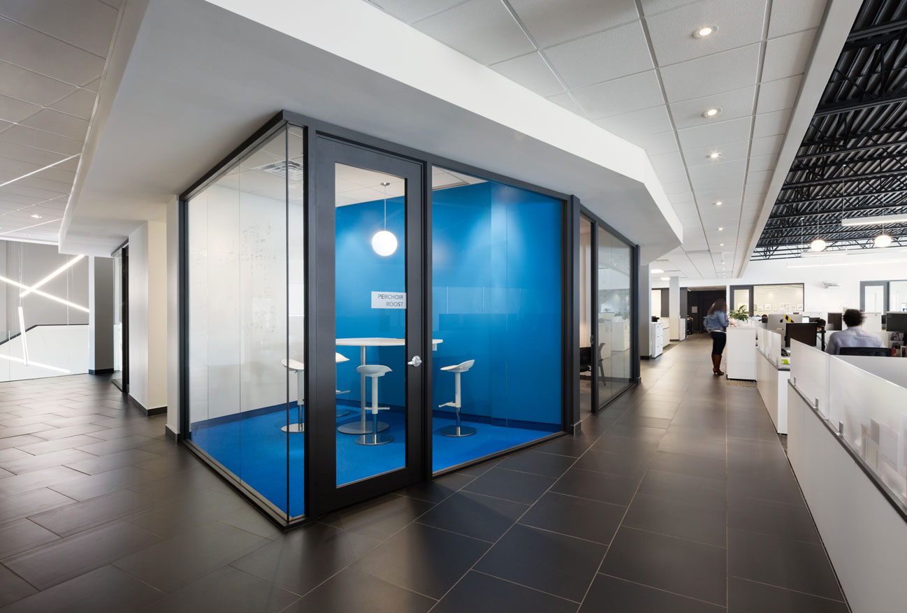 Glass cube meeting room with blue wall and floor with view of open corridor and person walking.