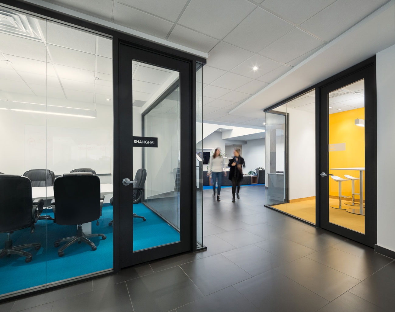 Corridor with glass cube meeting rooms on left and right, left with turquoise carpet, right with yellow wall and carpet.